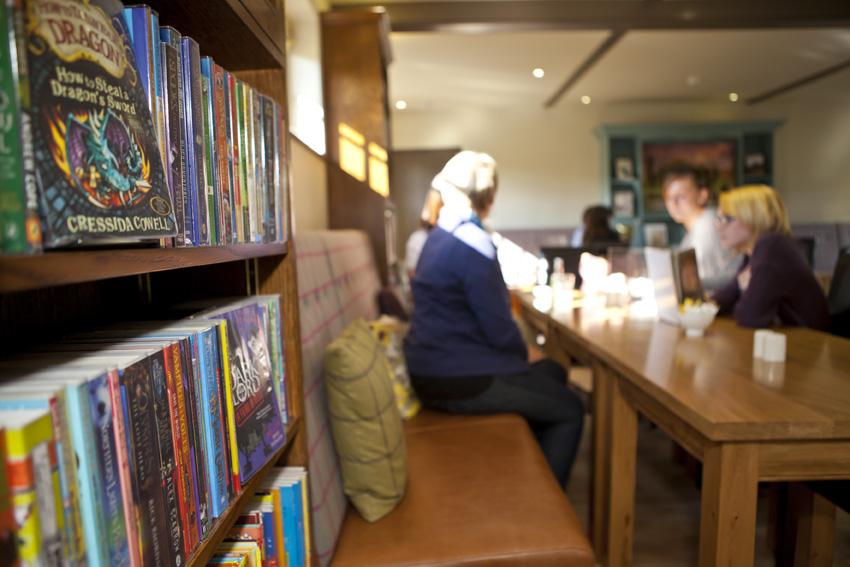 A group of customers sitting around a table at Rowntree Park Reading Cafe, with one half on padded benching and the others on dining room chairs.  In the foreground some paperback children's fiction is shelved in between seating areas.