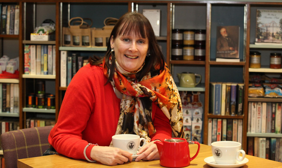 Fiona is a dark haired woman. She is smiling and wearing a bright red cardigan and print scarf. She is drinking a cup of tea and has bookshelves behind her