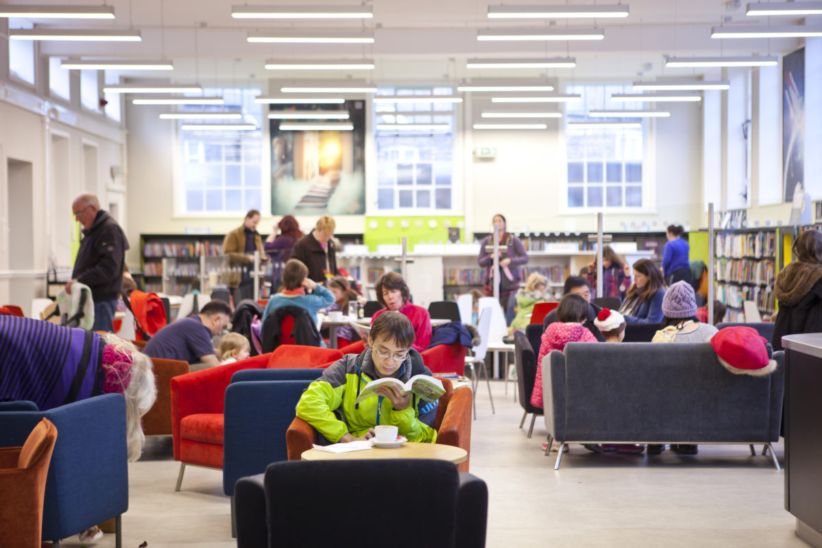 The café at York Explore library. A man in the foreground is sitting in a comfortable armchair, reading while he drinks a cup of coffee - he is wearing a lime green jacket and glasses. Behind him are groups of people sitting on sofas and parents with their children looking around the children's library.