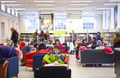 The café at York Explore library. A man in the foreground is sitting in a comfortable armchair,reading while he drinks a cup of coffee - he is wearing a lime green jacket and glasses. Behind him are groups of people sitting on sofas and parents with their children looking around the children's library.