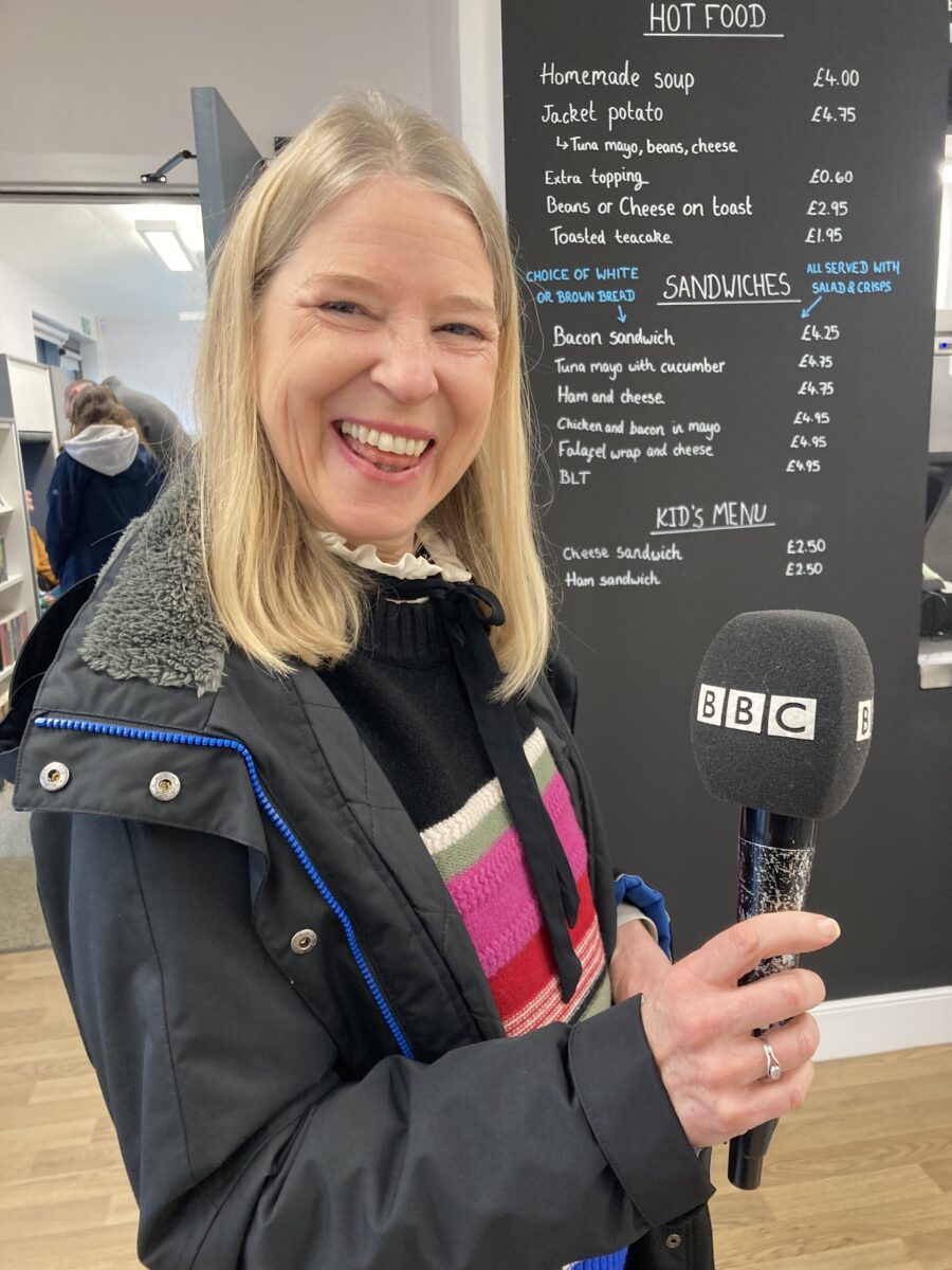 Cathy Killick from BBC Look North is holding a large microphone labelled "BBC". Cathy has blonde hair and is wearing a striped sweater and she is smiling.