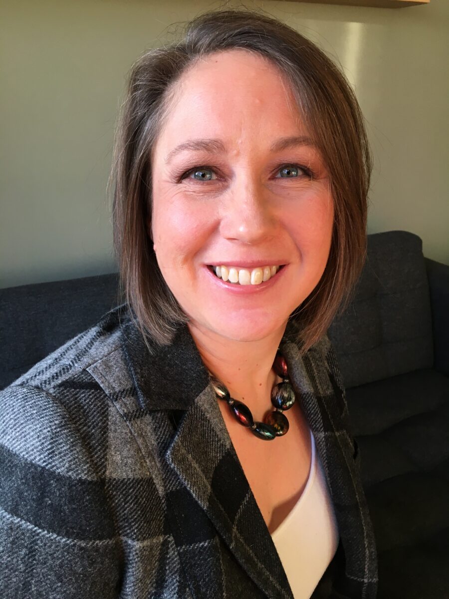 A dark-haired woman wearing a grey checked jacket, white top and necklace. She is smiling