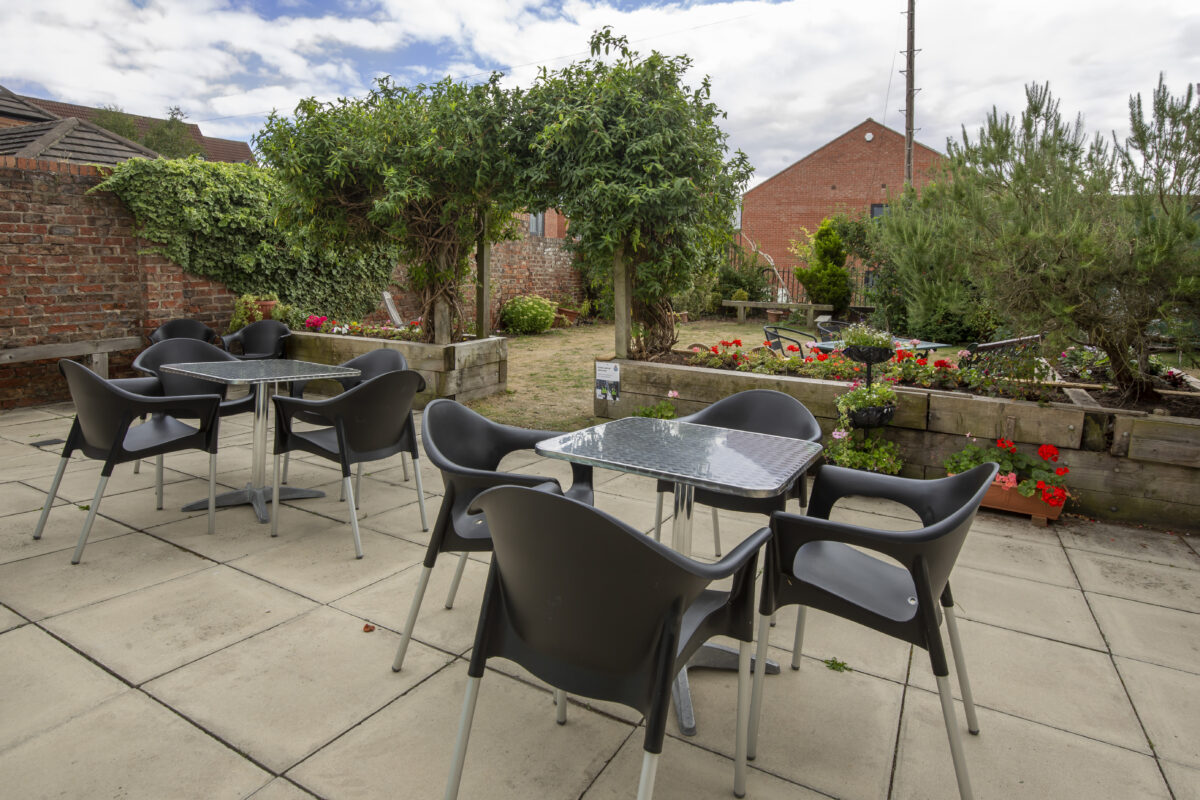 The garden at Acomb Explore set with cafe tables. The planters are colourful with red geraniums. 