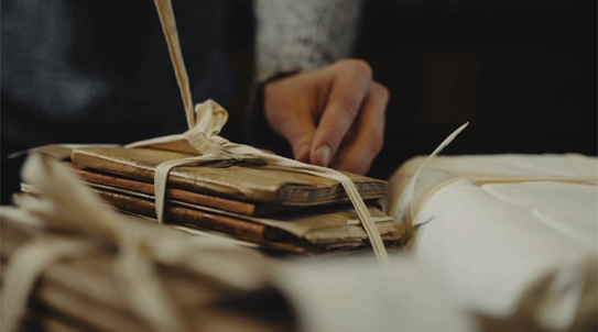 A person's hand, likely that of a researcher or historian, extends towards a stack of aged books bound together with twine. The books, with their faded covers and worn spines, suggest a collection of historical documents or personal writings. 