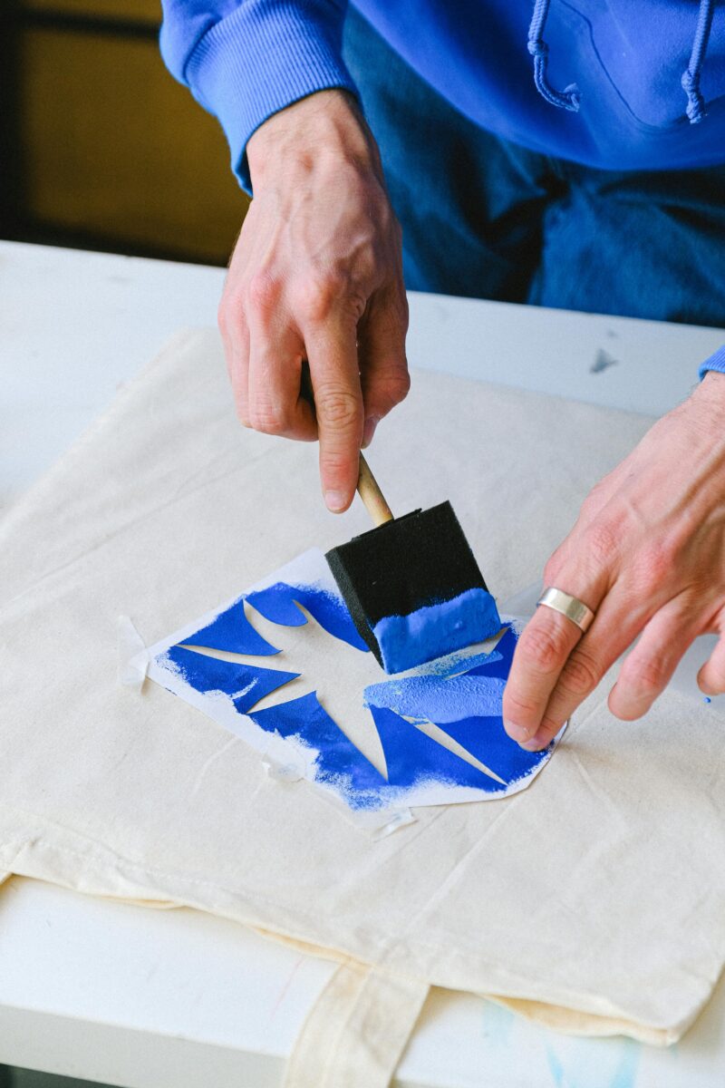 A close-up photo of a person's hands carefully applying blue paint with a brush to a stencil on a white canvas tote bag. 