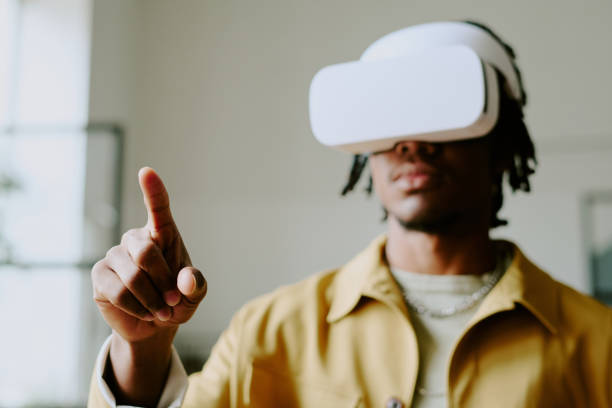 A person wearing a white virtual reality headset has their right index finger extended as if interacting with a digital environment. The individual is wearing a mustard yellow jacket and a silver chain necklace.