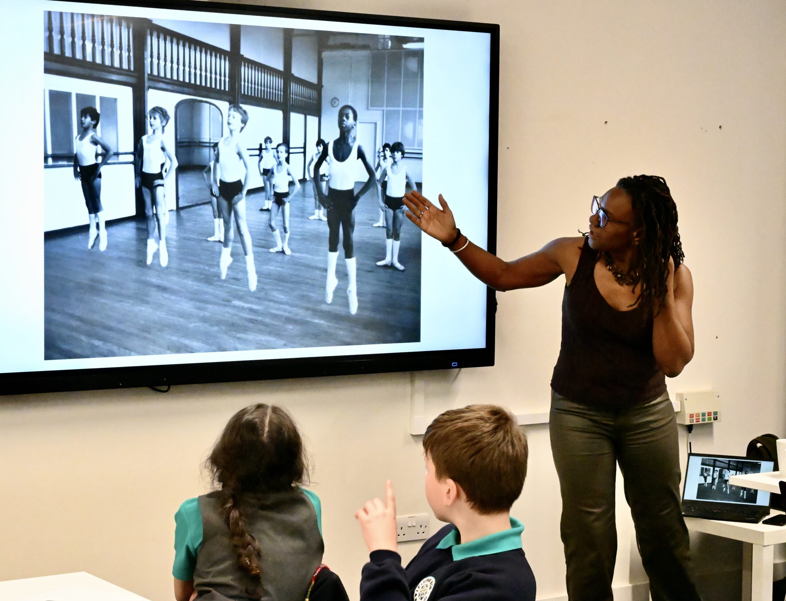 A teacher with dreadlocks and glasses points to a large screen displaying a black and white photograph of young ballet dancers in a studio. Two children sit at a desk in the foreground, listening to the teacher.