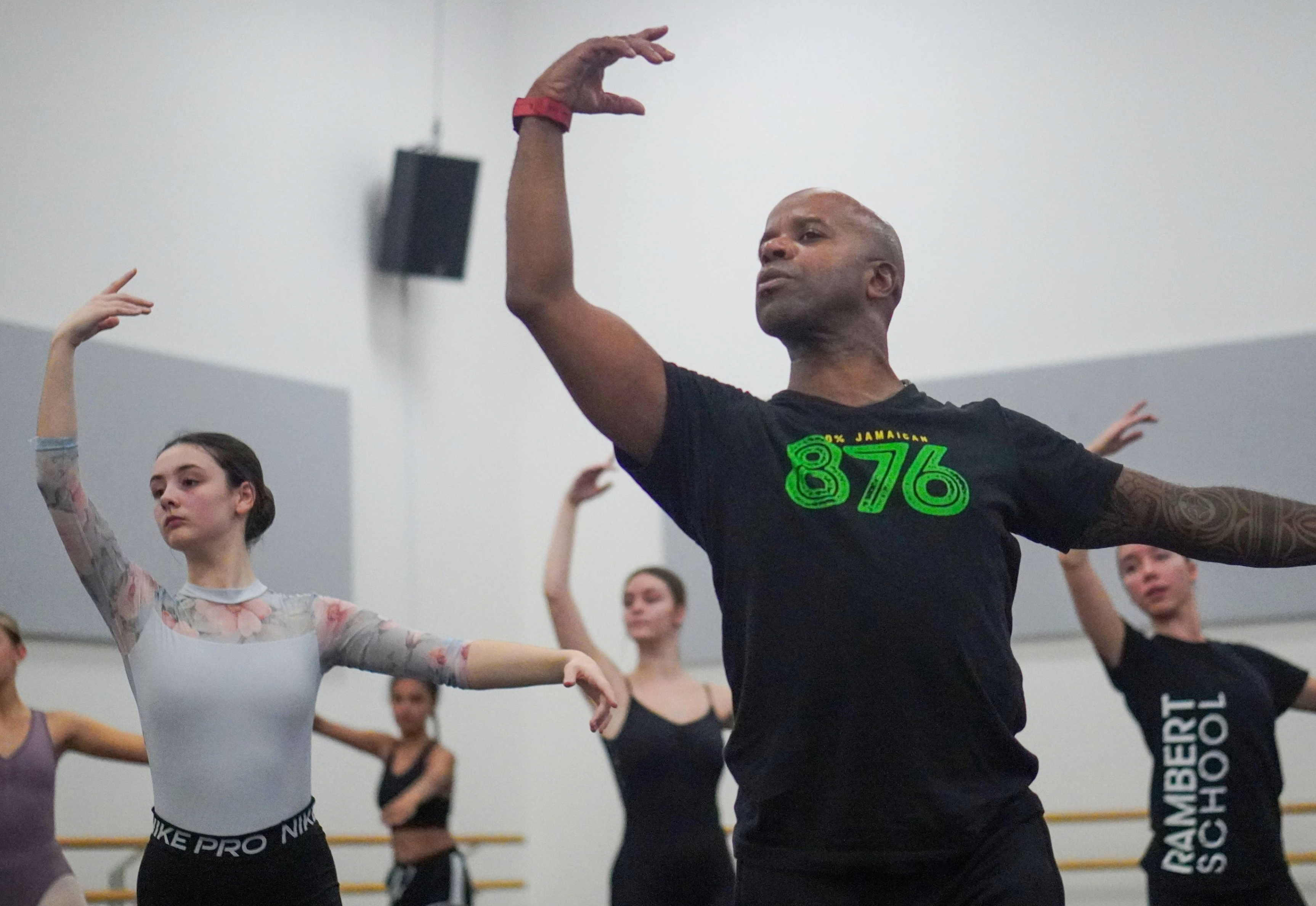 A male ballet teacher in a black t-shirt with green writing demonstrates a ballet pose with one arm raised and the other extended to the side. Several young female ballet students in leotards mirror his position in a bright studio.