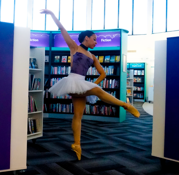 A ballet dancer in a purple leotard, white tutu, and gold pointe shoes gracefully poses en pointe in a library. Surrounded by bookshelves labelled "Fiction", she has one arm raised and the other extended to the side, showcasing a poised and elegant ballet position.