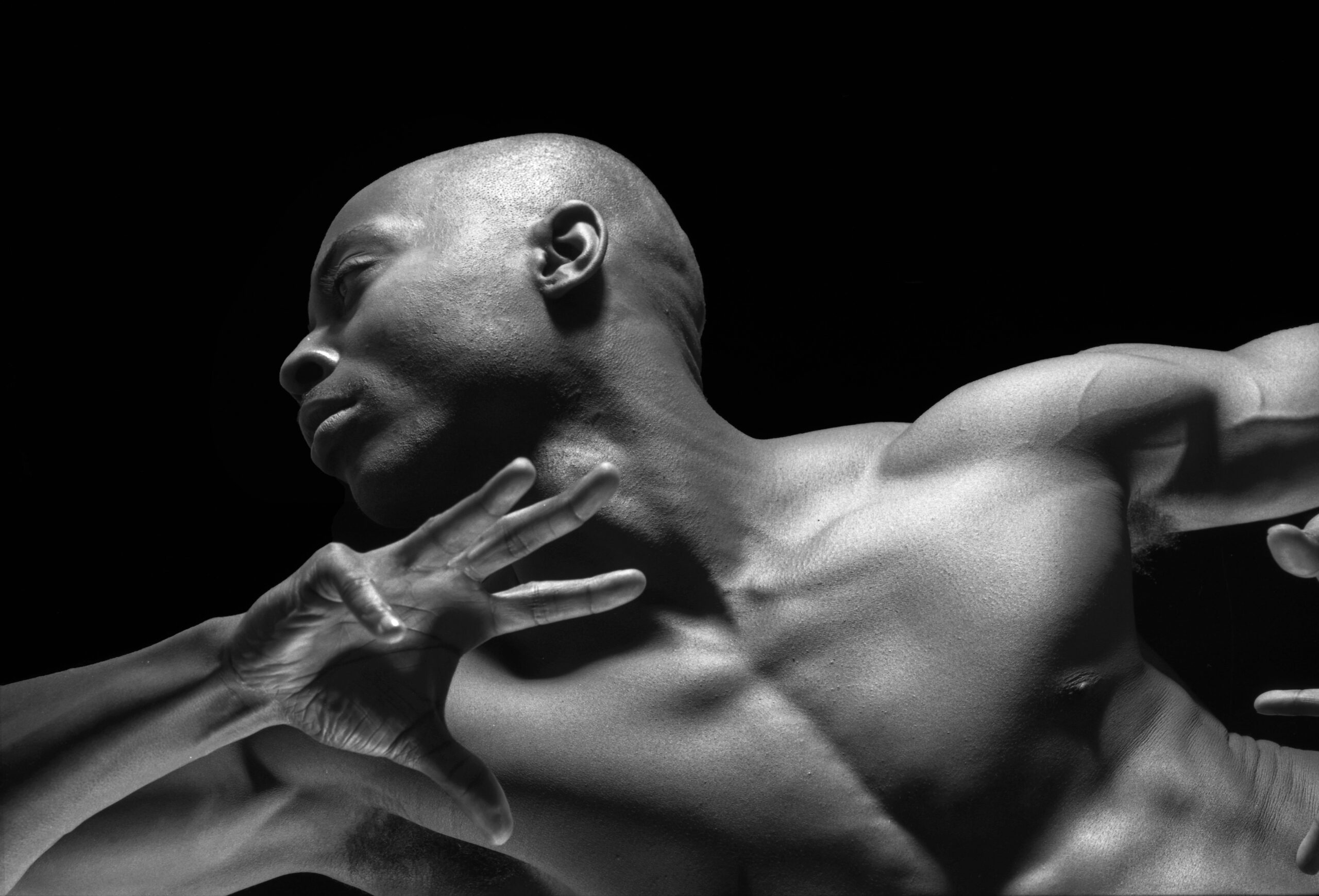 A close-up black and white photograph captures a muscular, bald ballet dancer in motion against a stark black background. His head is turned to the left, and his arms are extended with hands open, conveying a sense of dynamic movement and emotion.