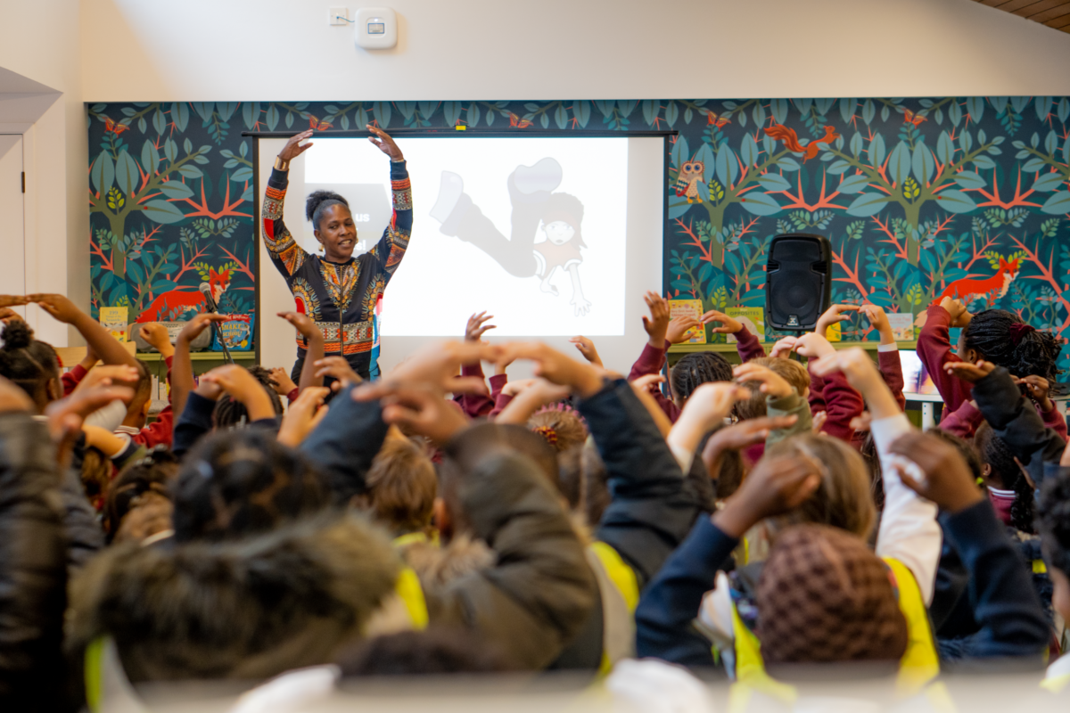 A teacher with arms raised leads a large group of children, who are also raising their arms, in a workshop. A projector screen behind the teacher displays a silhouette of a dancing figure against a backdrop of patterned wallpaper featuring animals and foliage.