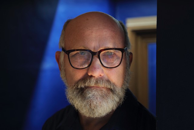 A photograph of Stephen Unwin looking directly at the camera. He has a beard, mustache and is wearing black glasses.