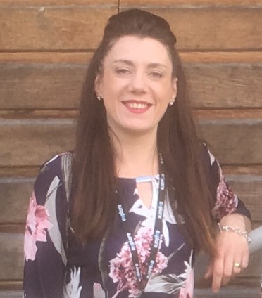 Katie is long black hair. She is wearing a patterned top and is smiling.